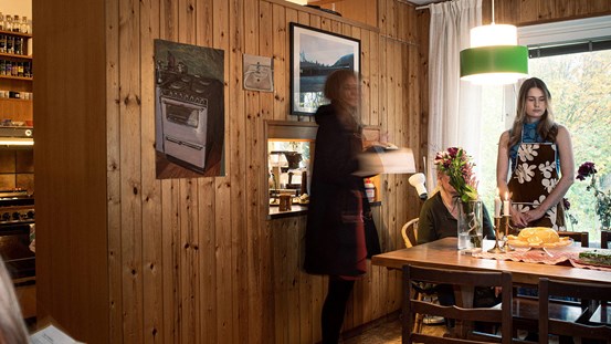 Dokumentation av köksinteriör där tre personer syns. På väggen hänger ett fotografi och två målningar.