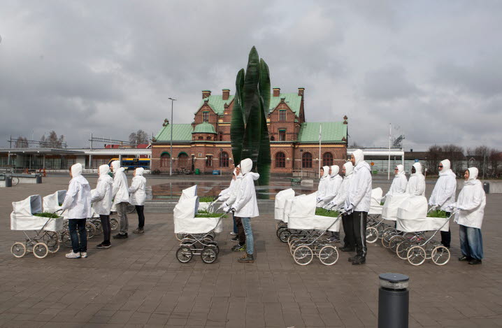 På ett torg står 16 personer i vitt med en vit barnvagn framför sig. I barnvagnarna växer gräs. Dokumentation från Gunilla Sambergs performance ”Convoy”.