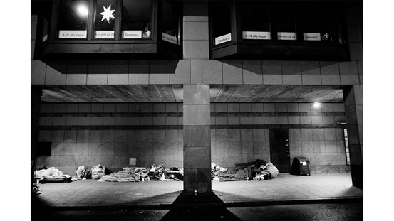 Åke Ericsons svartvita fotografi av romer på Olof Palmes gata i Stockholm. Under ett upplyst utomhustak ligger människor och sover.