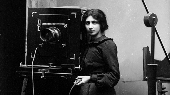 Svartvitt fotografi som visar en ung kvinna, Amalie Huczkowski Skogman, vid en lådkamera. I handen håller hon självutlösaren som tar fotografiet. Fotografiet är beskuret.