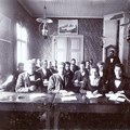 Studerande elever i en gammeldags lektionssal med kakelugn och en fotogenlampa i taket.