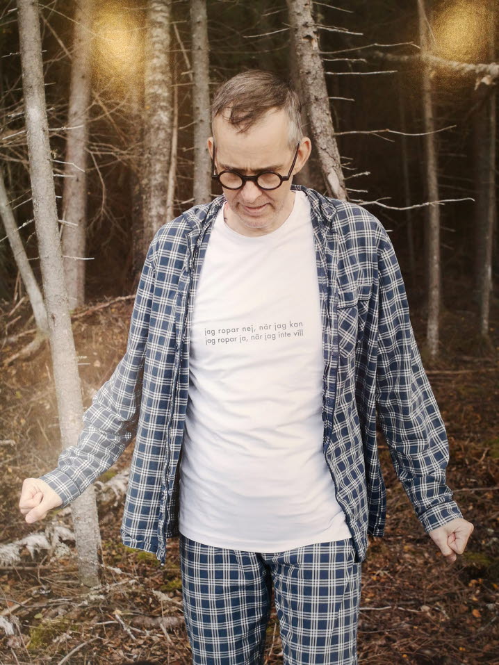 Färgfotografi av en man i blårutig skjorta och byxor och en vit t-shirt. I bakgrunden syns några träd. ©Micael Norberg/Bildupphovsrätt 2020.