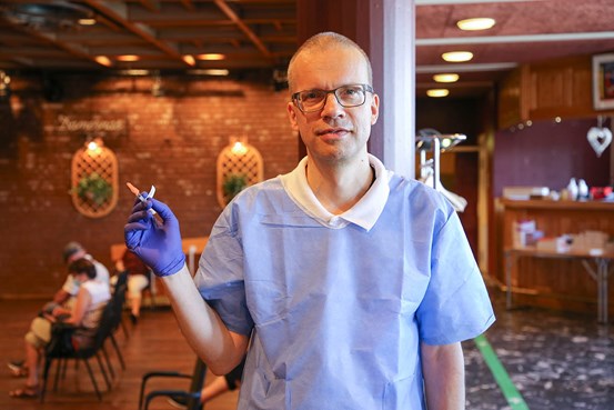 Benno Krachler i halvfigur, har blå syddsrock, håller upp en vaccinspruta med höger hand som han har en lila skyddshandske.