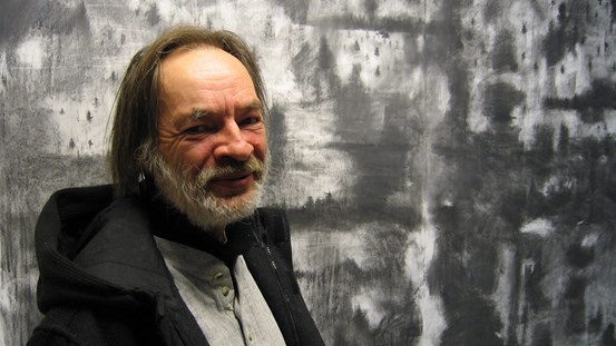 Porträttfoto av Christer Carlstedt. Man med skägg iklädd luvtröja, i bakgrunden en teckning i gråskala. 