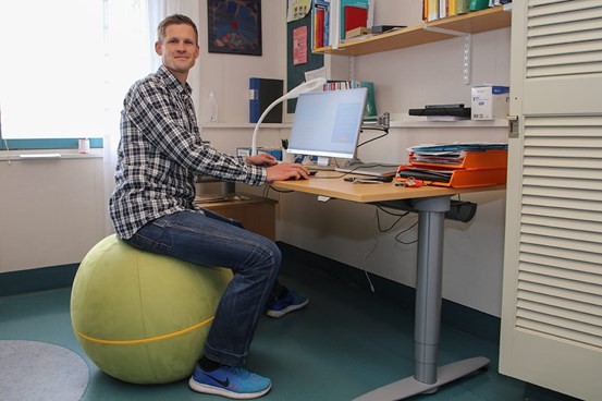 Daniel Carlsson sitter på en stor boll vid ett skrivbord där det står en dator. 