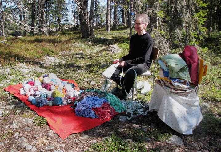 Håkan Bring sitter på en stol i naturen och klipper mattrasor. Färdigrullade nystan ligger på den röda filten vid hans fötter.