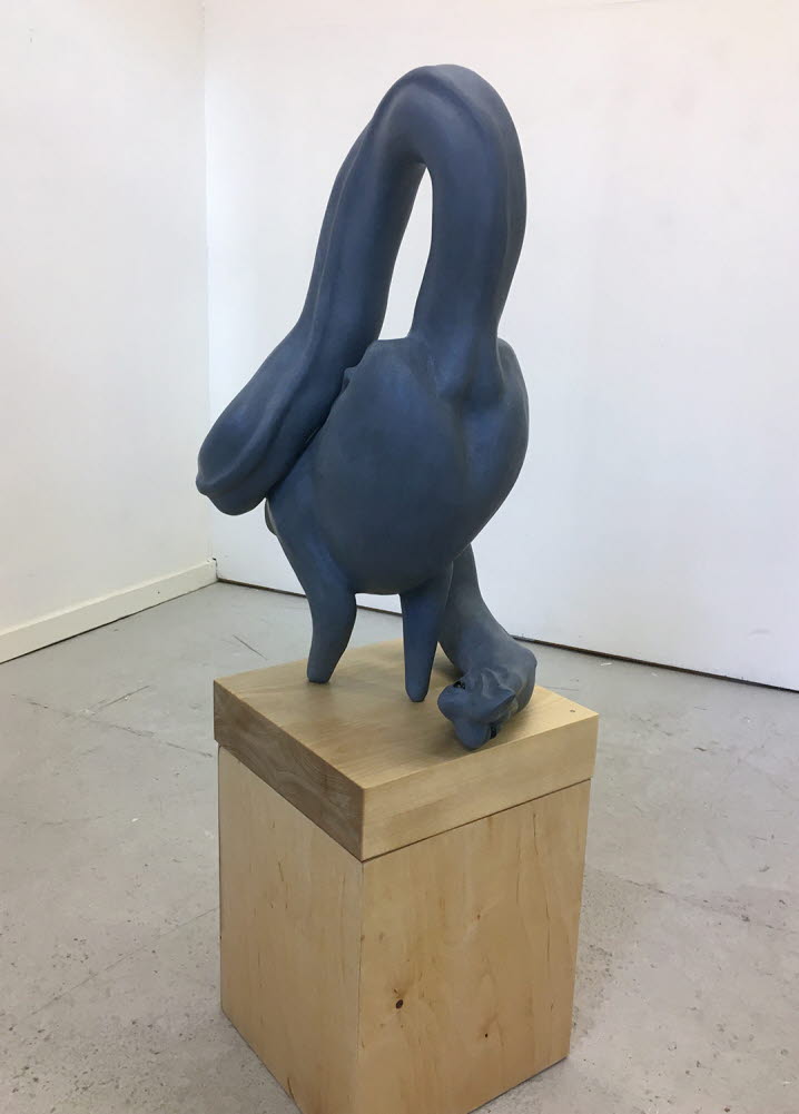 En gråblå skulptur, stående på ett podium, ensam i mitten av rummet, som representerar ett djur, ett monster, en sorts fågel, kanske en svan med en mycket lång och snirklig hals och ett huvud som ligger vid dess fötter. 