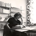 Kvinna med lockig 80-talsfrisyr som sitter i ett rum med bokhyllor och läser i en bok.