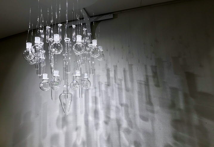 Skulptur av återbrukade objekt av Kerstin Hedström. Från taket hänger gamla laboratorieglas som bildar formen av en kristallkrona. På väggen syns skuggor och ljusreflektioner.