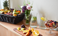 Bord med en fruktkorg, blommor i en vas, några karaffer med vatten, en godisskål och uppskuren apelsin på en skärbräda.