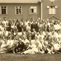 Personer i gammeldags kläder på en gräsmatta utanför en träbyggnad samlade för en gruppfotografering.