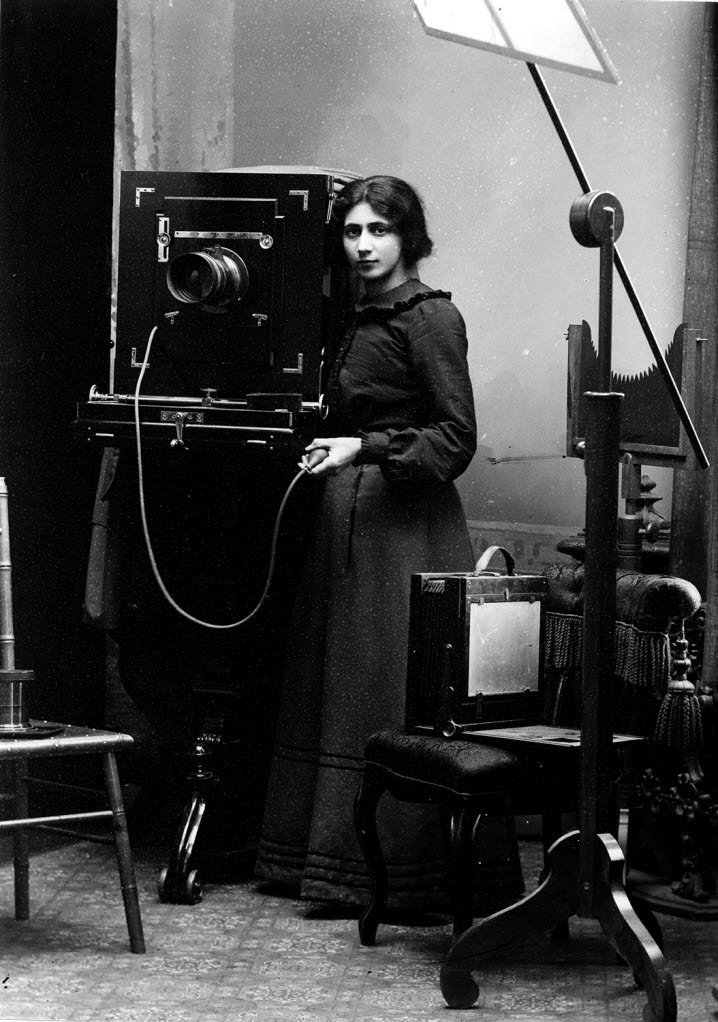 Svartvitt fotografi som visar en ung kvinna, Amalie Huczkowski Skogman, vid en lådkamera. I handen håller hon självutlösaren som tar fotografiet.