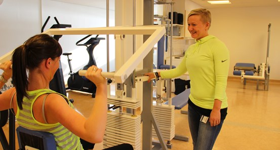 En person som sitter med ryggen mot kameran och lyfter vikter och en person som står med ansiktet mot kameran och ler mot personen som tränar.