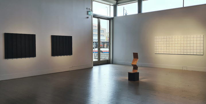 Utställningsvy från konsthallen i Luleå. På vänstra väggen hänger två närmast svarta verk som påminner om husfasader. På högra väggen hänger ett vitt verk som påminner som klinkers. På golvet står en skulptur av trä.