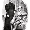 Skallig sittande man i ljus kostym och en stående kvinna i mörk klänning som håller mannen på axeln.