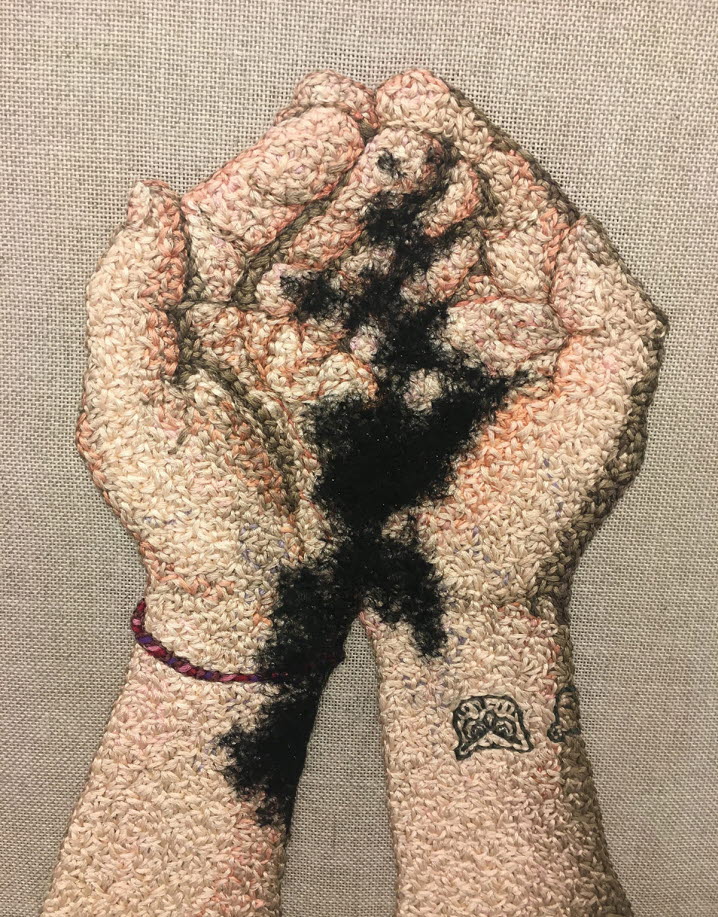 Ett inramat broderi som representerar sammanhållna händer med öppna handflator, en tatuering med en upp och nervänd uggla pryder höger armen, ett rött svart armband sitter på vänstra handleden, hudfärgat garn, samt svart yvigt garn som sprider sig mellan händerna och ner på armarna.