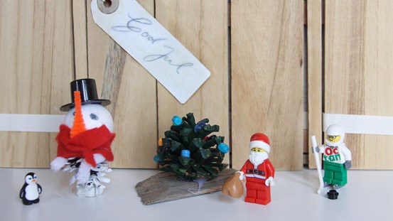 Ett foto av hemgjorda julpynt, en leksakspingvin, en legotomte och en legofigur ståendes framför en vägg. På väggen sitter en etikett med texten god jul.