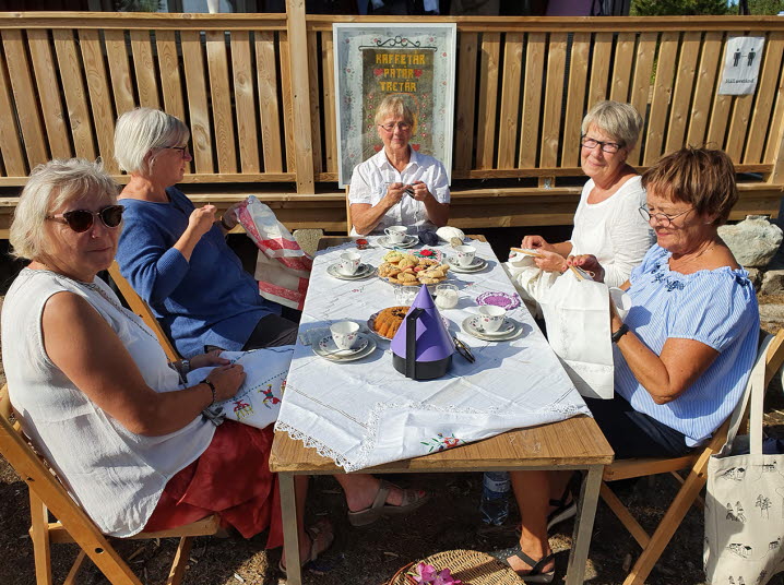 Fem handarbetande kvinnor sitter runt ett bord uppdukat med kaffe och kakor. 