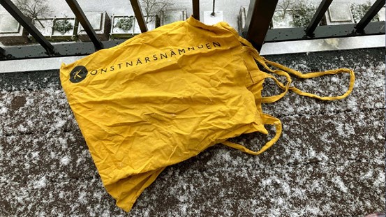 Foto, gul tygkasse med Konstnärsnämndens logotyp tryckt längs ena sidan. Kassen ligger på ett golv intill balkongräcke. Ett tunt lager snö har lagt sig på golvet. 