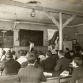 Elever och en undervisande lärare i en lektionssal med två griffeltavlor och några fotogenlampor i taket. 