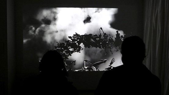 Dokumentation av Magnus Wallins animerade videoverket ”Elements”. Svartvit animation projiceras mot en vägg, föreställande skelett som svävar mot en himmel. Två personer tittar på videoverket och ses som svarta silhuetter i förgrunden.