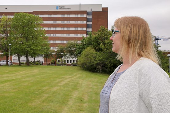 Maria frank står utanför sjukhuset i Sundsvall. Hon står framför en gräsmatta och tittar mot sjukhuset. Det är sommar och gröna träd syns på bilden.