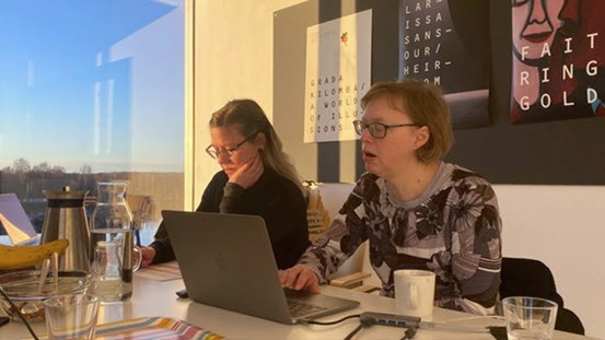 Lisa Lundström och Fanny Carinasdotter sitter vid ett skrivbord. En dator står på bordet. Solen skiner in genom ett stort fönster i bakgrunden där man även ser en blå himmel.