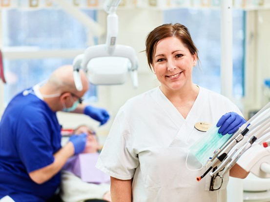 Tandsköterska i ett behandlingsrum, suddig bakgrund där en tandläkare behandlar en patient.