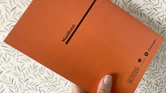 En hand håller en orange bok med titeln ”Manifest”.