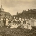 Personer klädda i gammeldags kläder i sommartid samlade för en gruppfotografering framför en träbyggnad. 