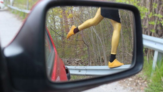 Foto av backspegel på bil. Däri speglas benen på en person ståendes på ett ben på ett vägräcke. 