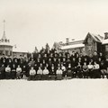 Elever i gammeldags kläder framför två träbyggnader i vintertid.