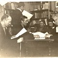 Personer i mörka kostymer som läser tidningar i ett rum med bokhyllor.