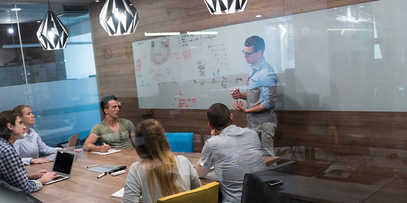 Personer i mötesrum lyssnar på en kollega som presenterar anteckningar på en stor whiteboard.