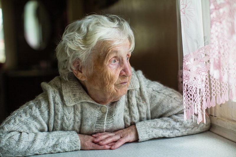 Äldre kvinna tittar ut genom ett fönster, ser ledsen ut