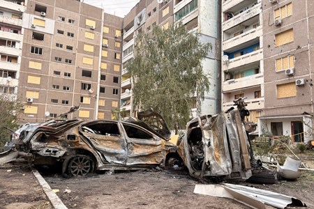 Utbrända bilar framför flerfamiljshus med skadade fasader och igenspikade fönster efter ett bombnedslag
