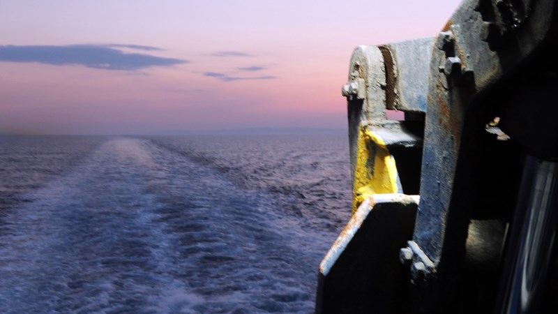 Vy från ett fartyg där aktersvallet syns i vattnet mot en rosa‑blå himmel, med en del av fartygssidan i förgrunden.