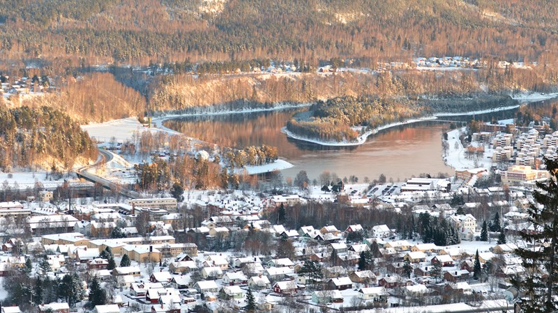 Snötäckt stad belägen vid en älv, omgiven av skogsklädda höjder i vinterlandskap.