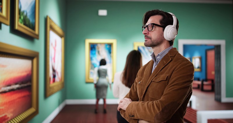 En man med hörlurar och glasögon står med armarna i kors och betraktar konstverk i ett museum. 