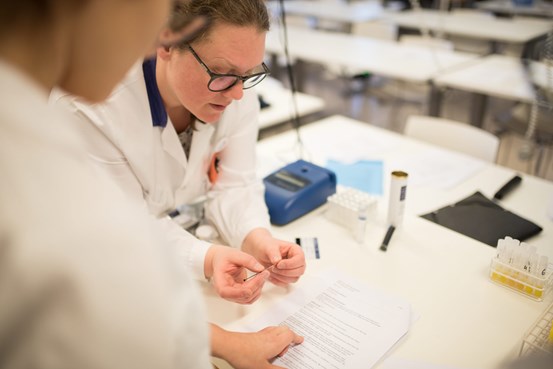 Två kvinnor i vita rockar. Den ena syns bakifrån och vi ser att hon läser ett dokumentdär hon håller sitt finger vid en textrad. Den andra lutar sig över bordet och håller i en smal remsa av något hon verkar studera närmare. 