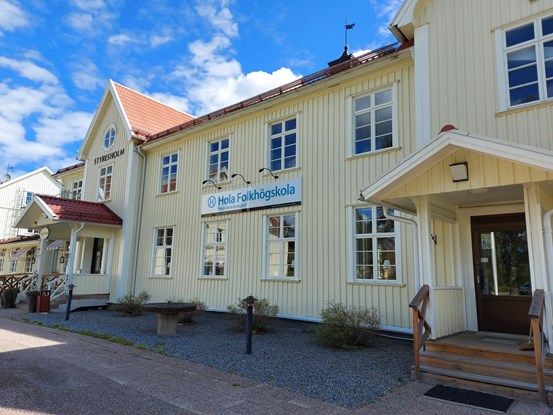 Skolans ljusgula fasad med skylten Hola folkhögskola mot en blå himmel.