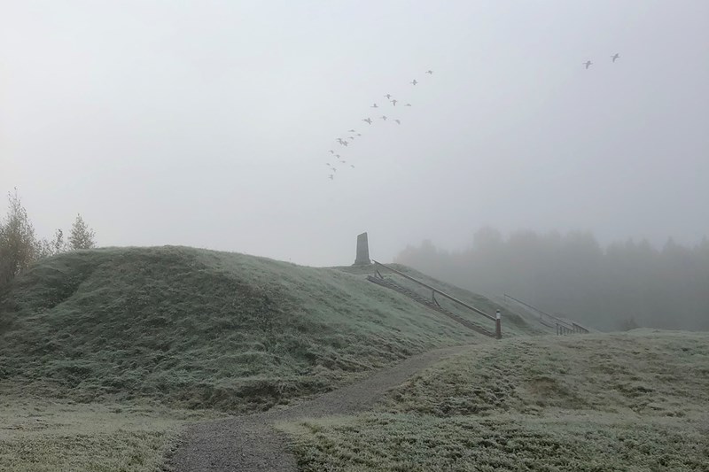 En kulle med ett stenmonument omringad av dimma och en flock fåglar på himmelen.