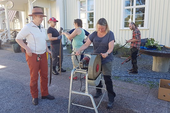 Sex personer håller i redskap och vässar en lie framför en gul byggnad