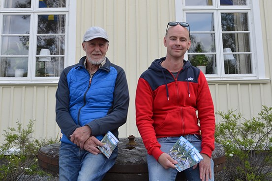 Två män står framför Hola folkhögskola med broschyrer i handen och tittar in i kameran.