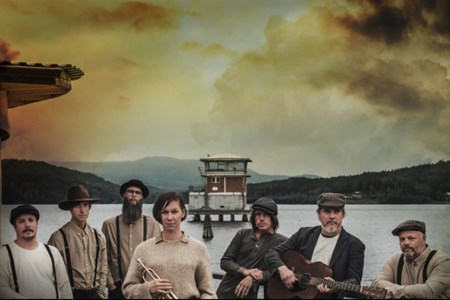 Sju musiker står och tittar in i kameran framför ett hav och bergslandskap.