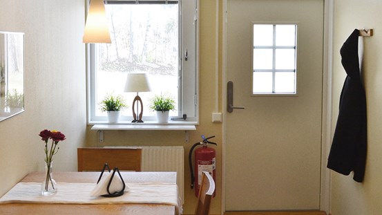 Köksbordet med två stolar, jacka som hänger på väggen, dörr till lägenheten samt fönster. 