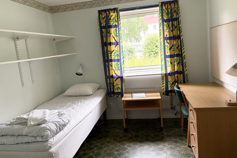 Ett rum med en säng, ett skrivbord, hylla och fönster med gardiner.