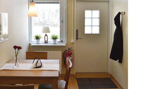 Köksbord med två stolar, jacka som hänger på väggen, dörr till lägenheten samt fönster.