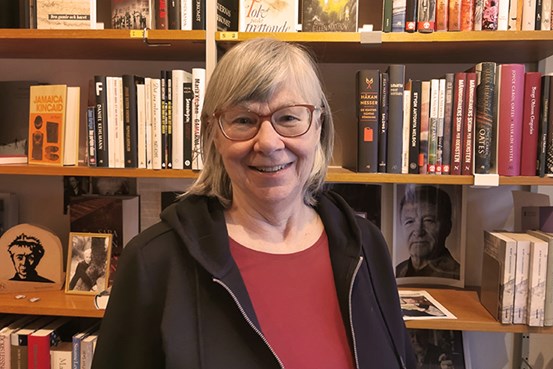 En kvinna i glasögon står glatt och tittar in i kameran framför en bokhylla full med böcker.