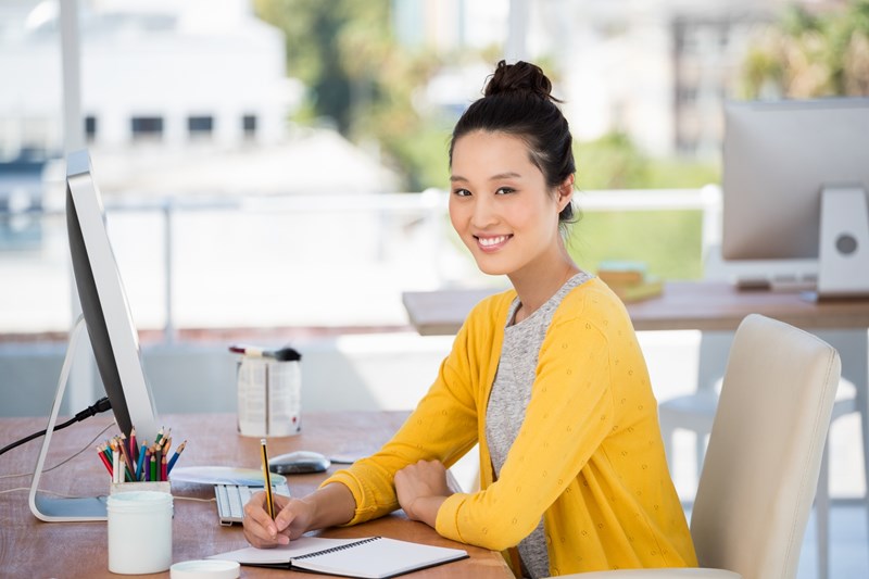 En kvinna i gul tröja sitter vid ett skrivbord med en dator och skriver med en penna på ett papper.